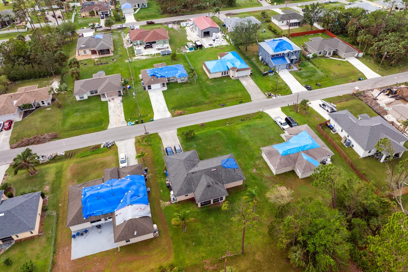 An aerial view of a suburban street where several houses have blue tarpaulins covering storm-damaged roofs
