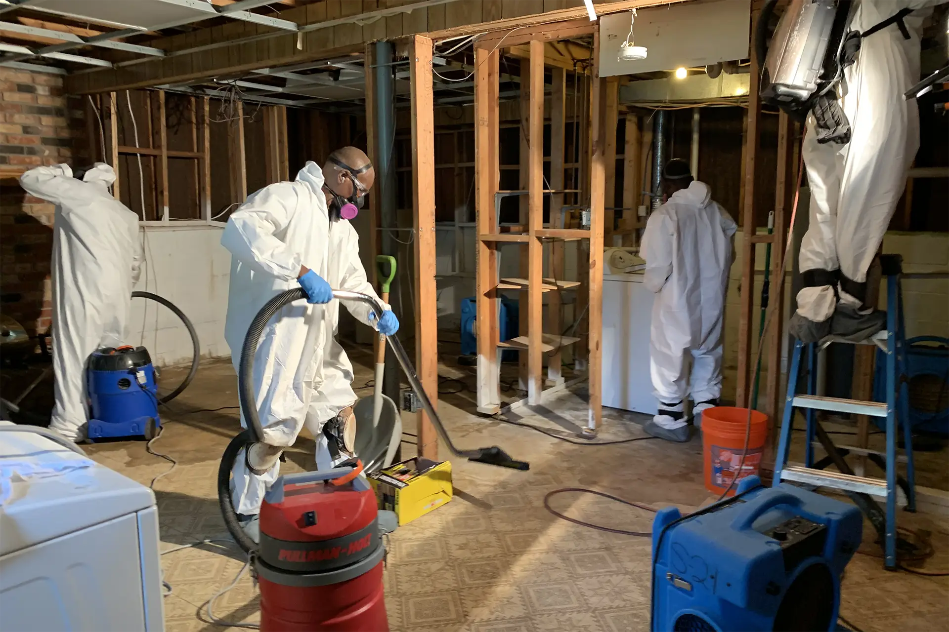 Restoration technicians in protective suits and respirators running drying equipment in a gutted basement