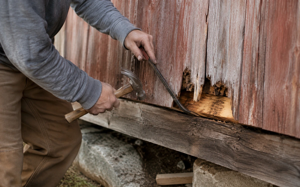 A worker prying rotted siding and sill boards from a weathered wood structure — restoration and radon insurance in Illinois