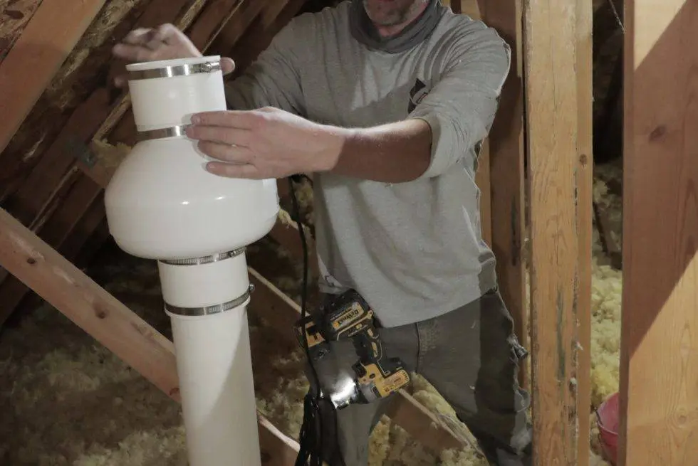 A technician assembling a radon exhaust fan and vent pipe in an attic with a cordless drill — radon mitigation insurance in Ohio