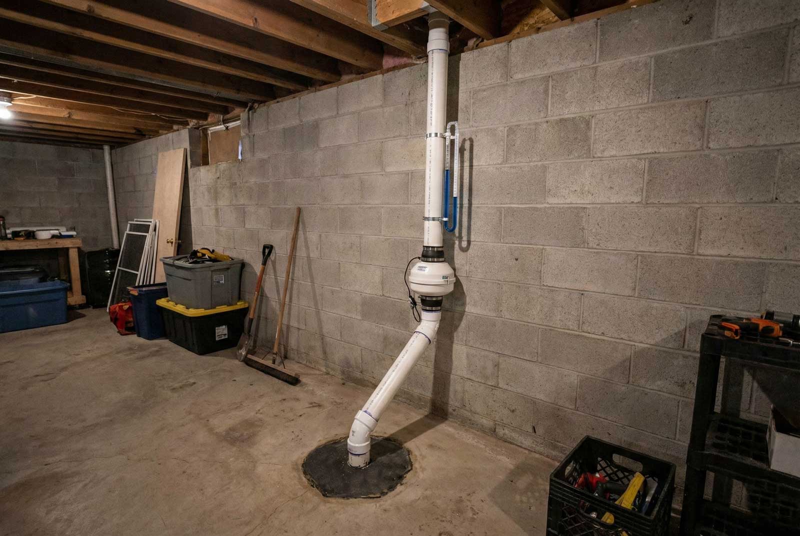A basement radon mitigation system with a suction pipe, an inline fan, and a U-tube manometer on a block wall — radon mitigation insurance in New York