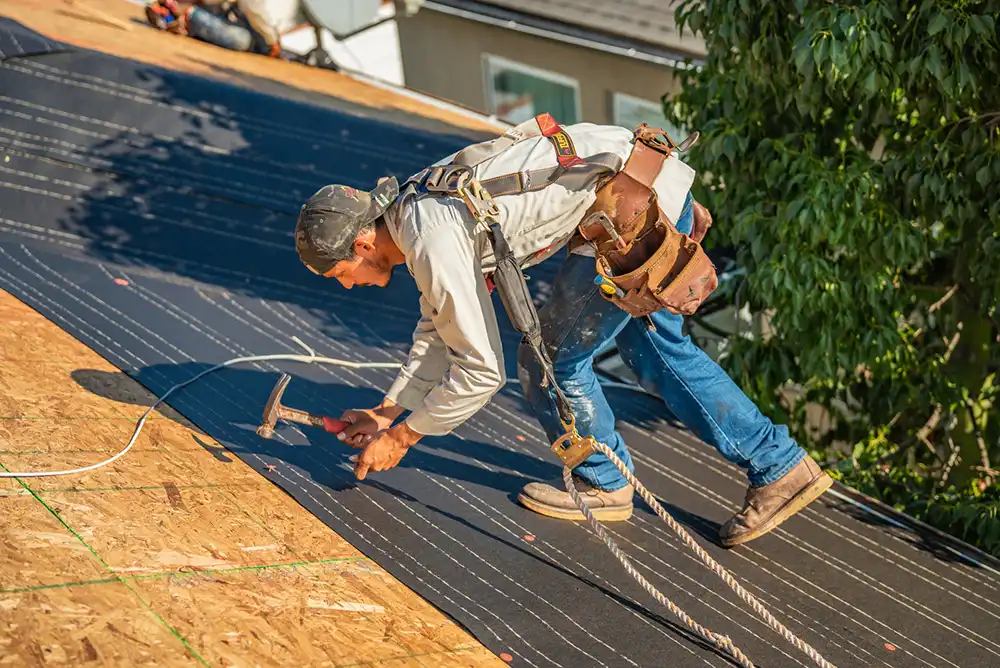 A worker in a fall-protection harness nailing underlayment across a sloped roof deck — restoration contractor insurance in Ohio